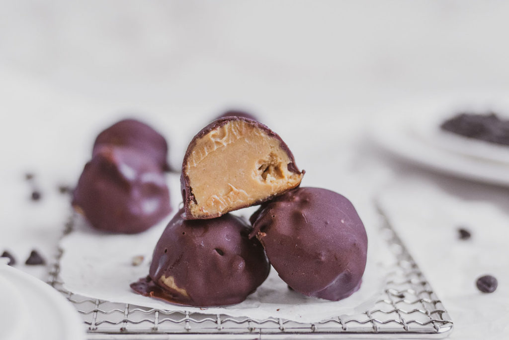 Keto Chocolate Peanut Butter Fat Bombs. Chocolate covered peanut butter balls on a white surface and wore wrack. 