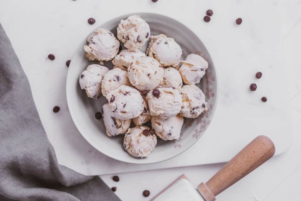Low carb keto cookie dough fat bombs in a white bowl on a white surface with chocolate chips scattered around and a grey napkin and rolling pin on the side.