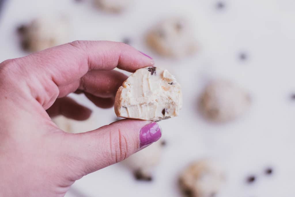 A hand holding a keto cookie dough fat bomb with a bite taken out. Low carb cookie dough fat bombs on a white surface in the background.