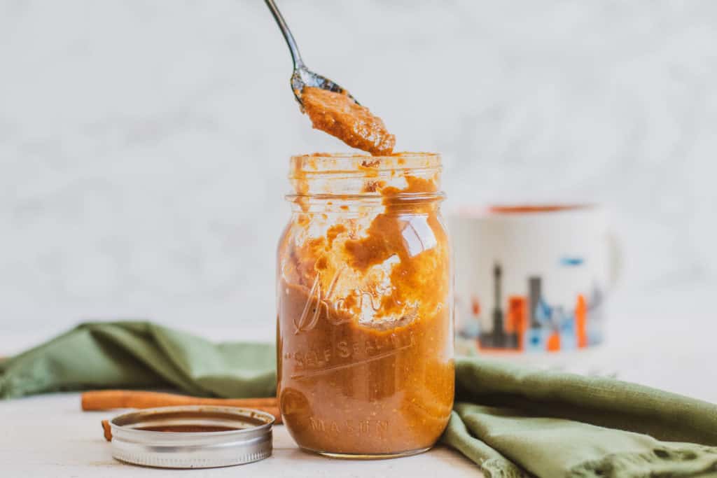 Low carb Pumpkin Spice Syrup in a glass jar, and a spoonful above, with cinnamon sticks on the side and a green napkin in the back.