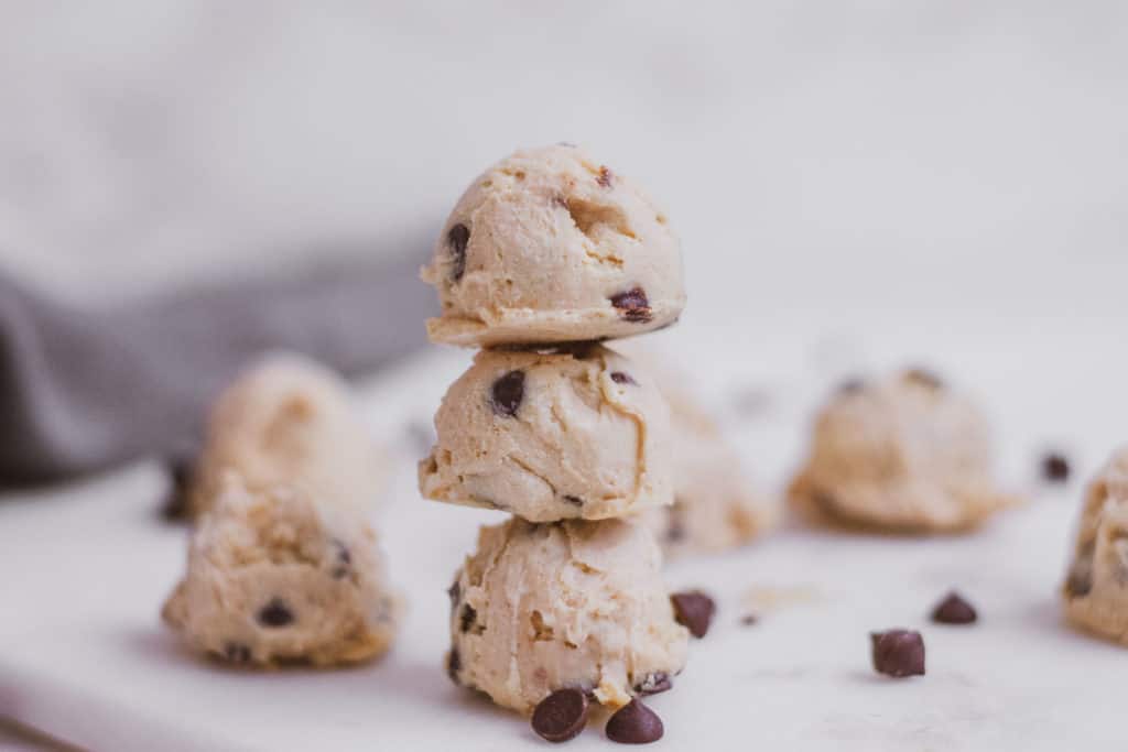 Low carb keto cookie dough fat bombs on a white surface stacked on top of each other with chocolate chips scattered around.