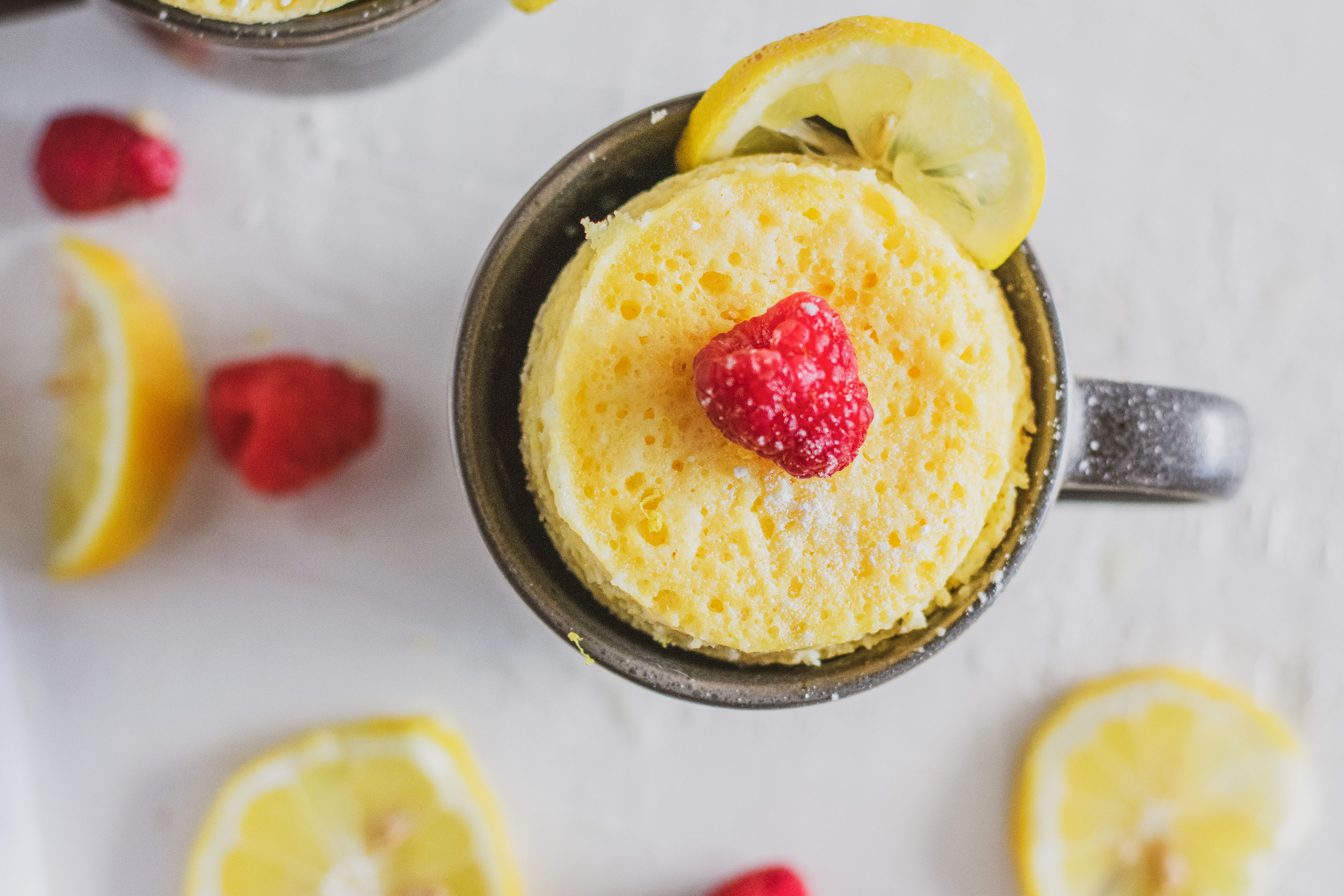 Low carb lemon mug cakes in a grey mug with raspberries and lemon slices on a white surface.
