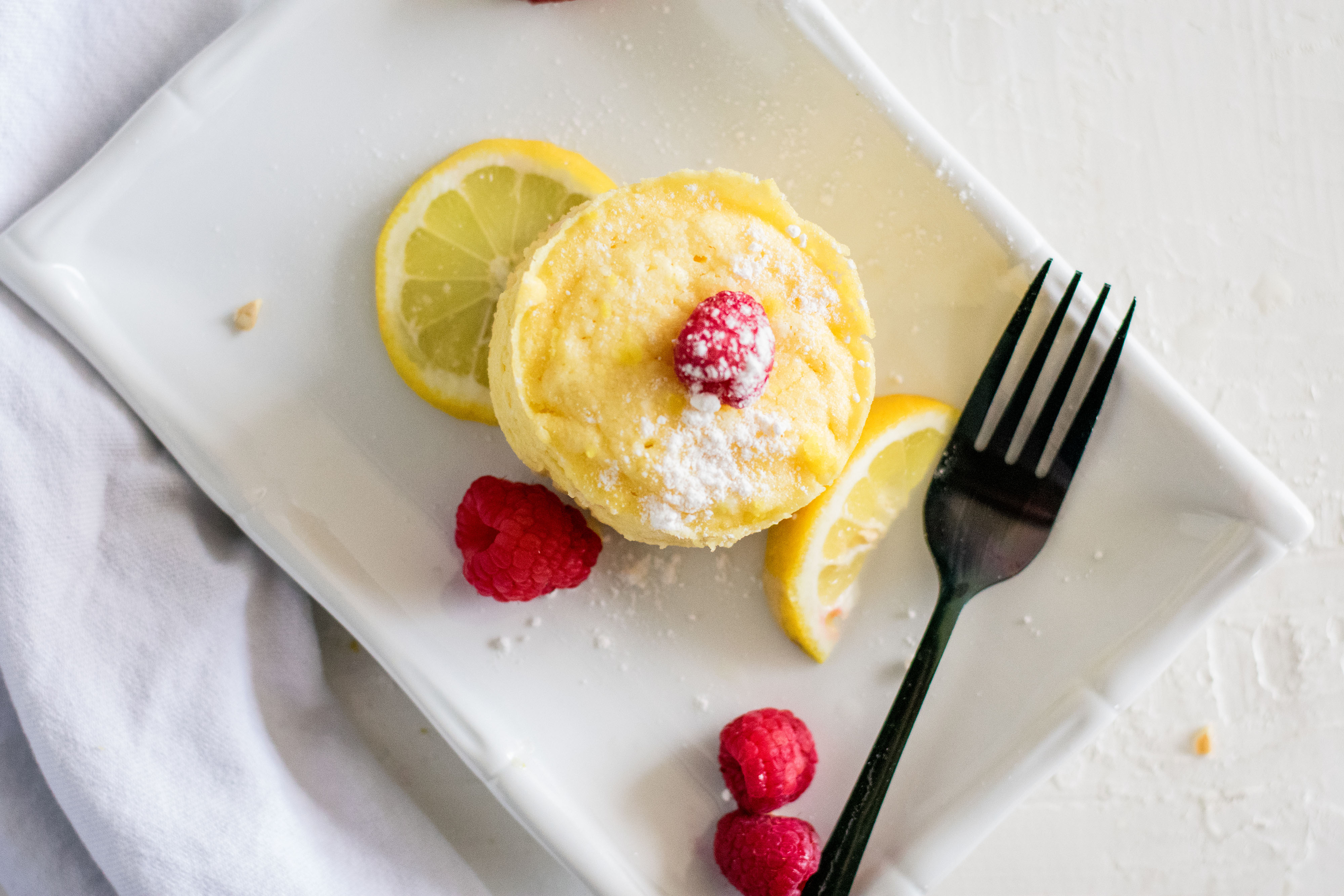 Low carb lemon mug cakes on a white plate with raspberries and lemon slices on a white surface.