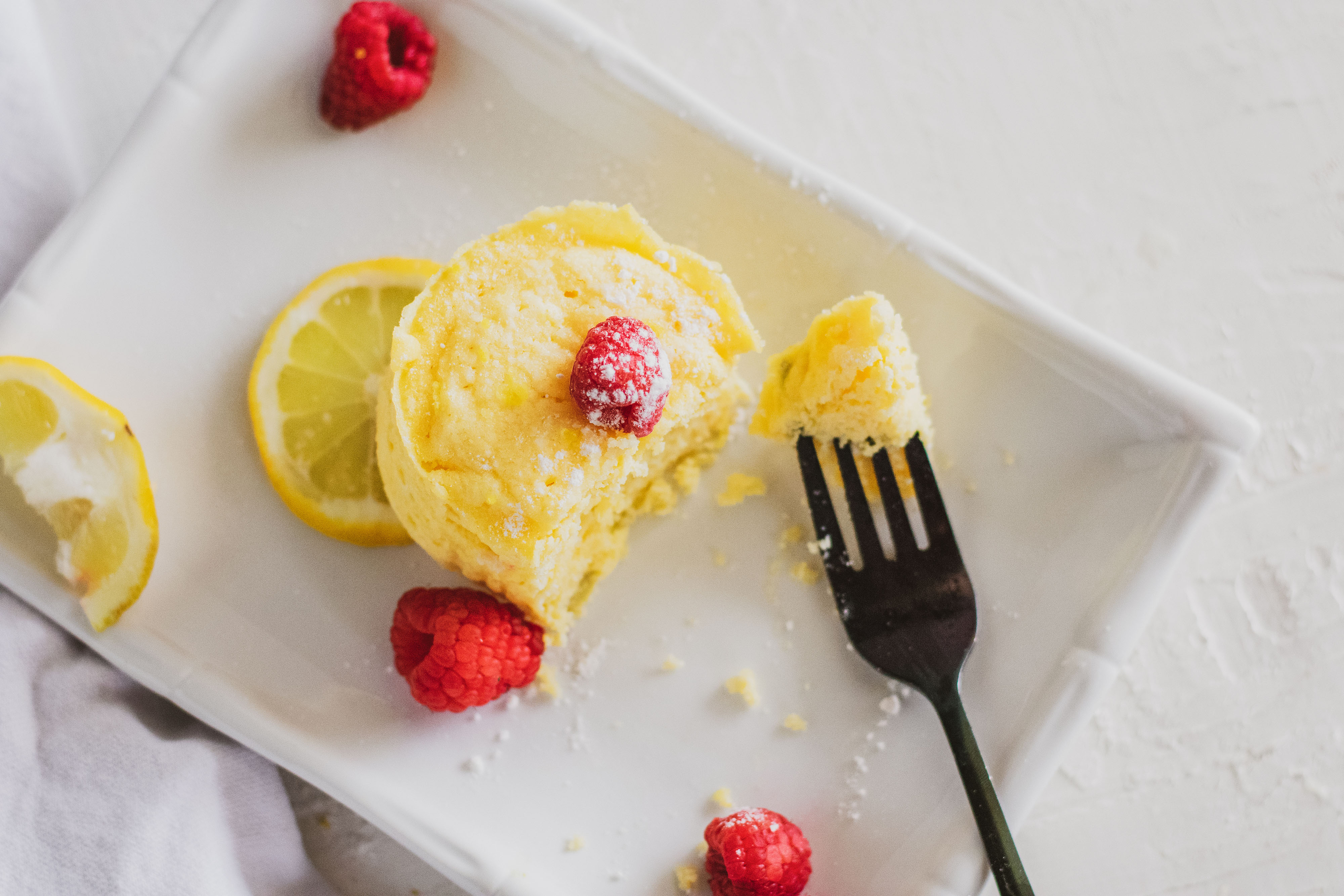 Low carb lemon mug cakes on a white plate with raspberries and lemon slices on a white surface.