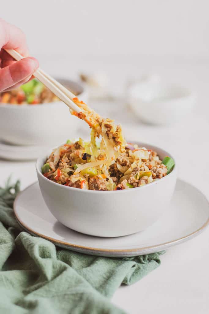 Keto Egg Roll in a Bowl with green onions and sriracha drizzled on top in a white bowl on a white plate on a green napkin with chop sticks picking up a bite