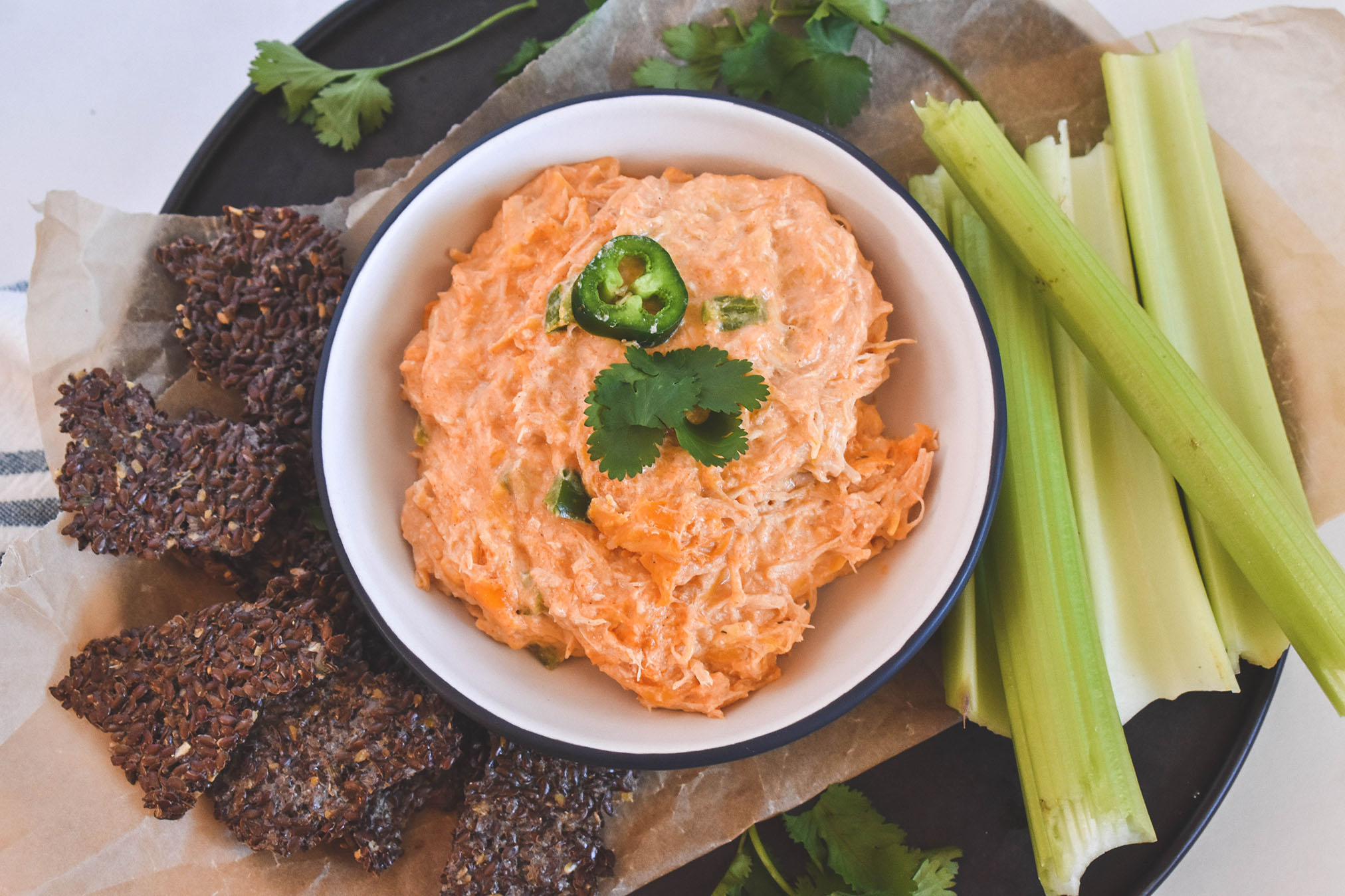 Buffalo Chicken dip in a white bowl sitting on parchment paper with celery sticks and flax crackers on the side.