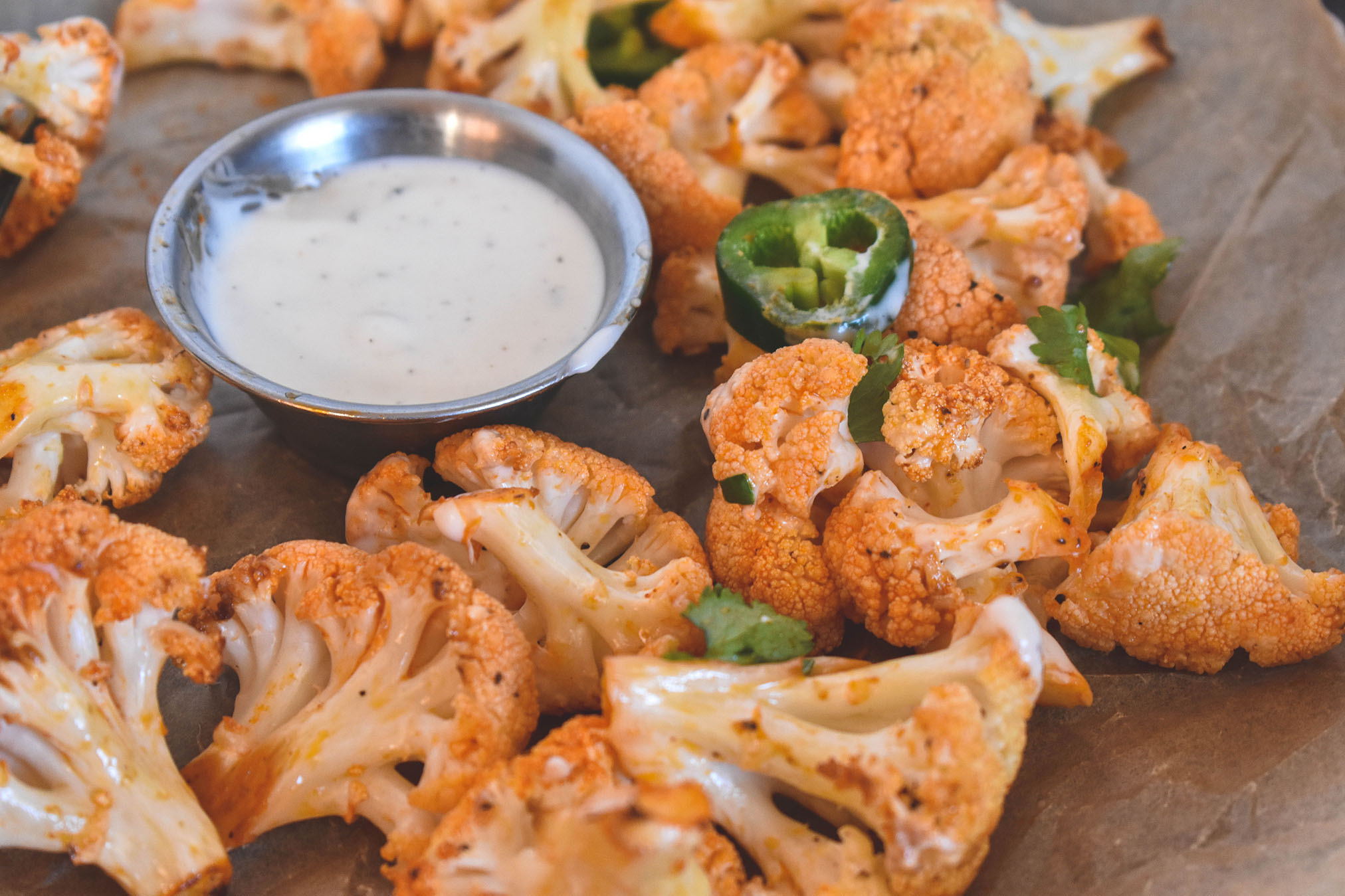 Buffalo sauce covered cauliflower bites cut up laying on parchment paper with a side of ranch and topped with jalapenos.