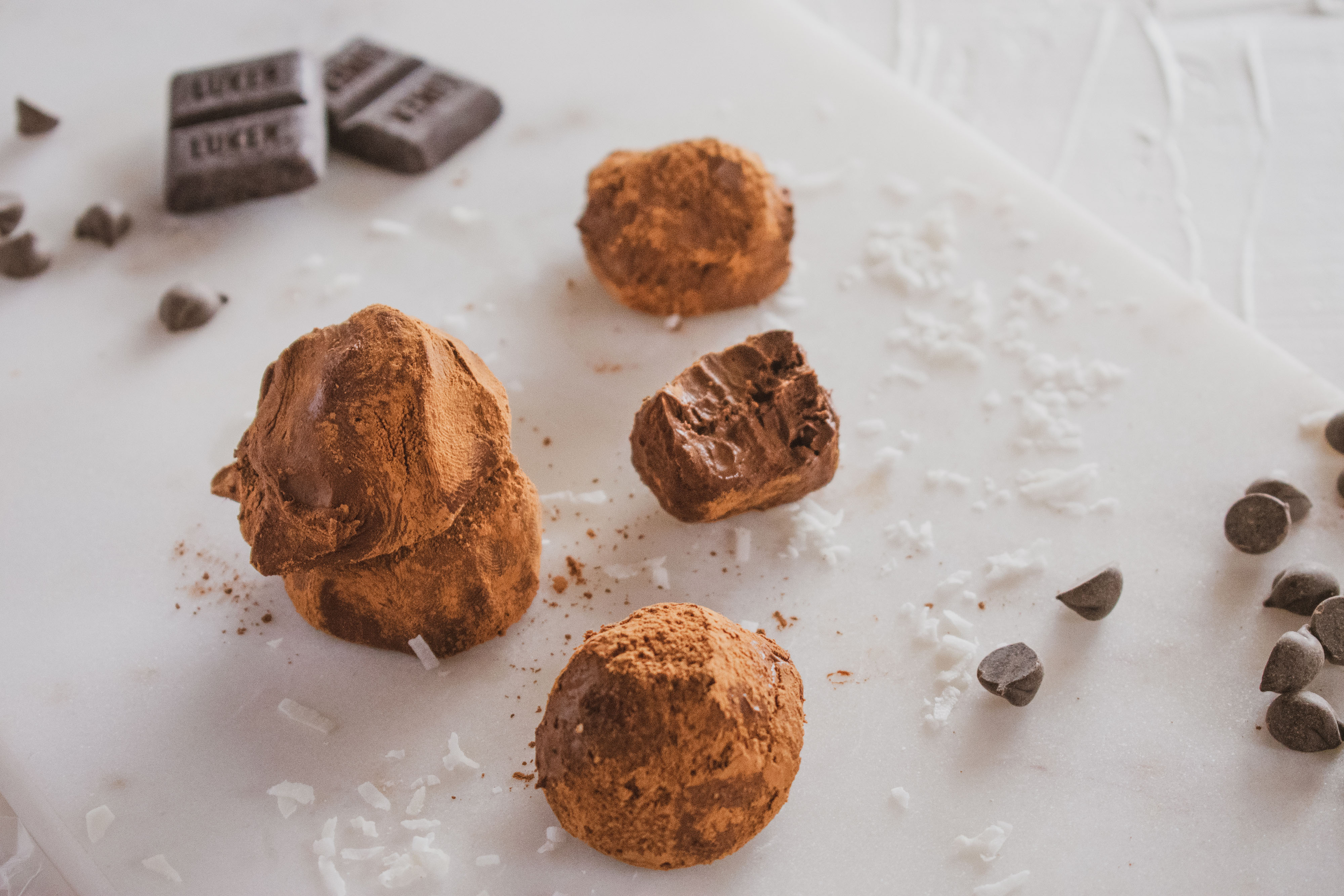 Cocoa powder covered keto chocolate truffles. Set on a white surface with chocolate chips and with a bite taken out.