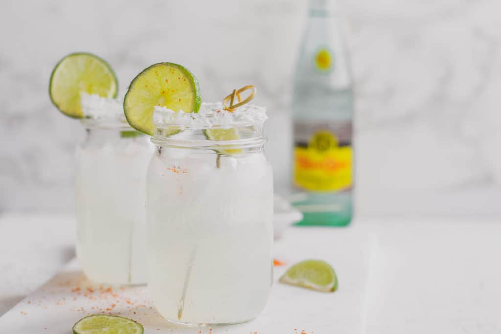 Keto margarita in a mason jar with salt and a sliced lime on the rim on a white surface with a topo chico on the side.