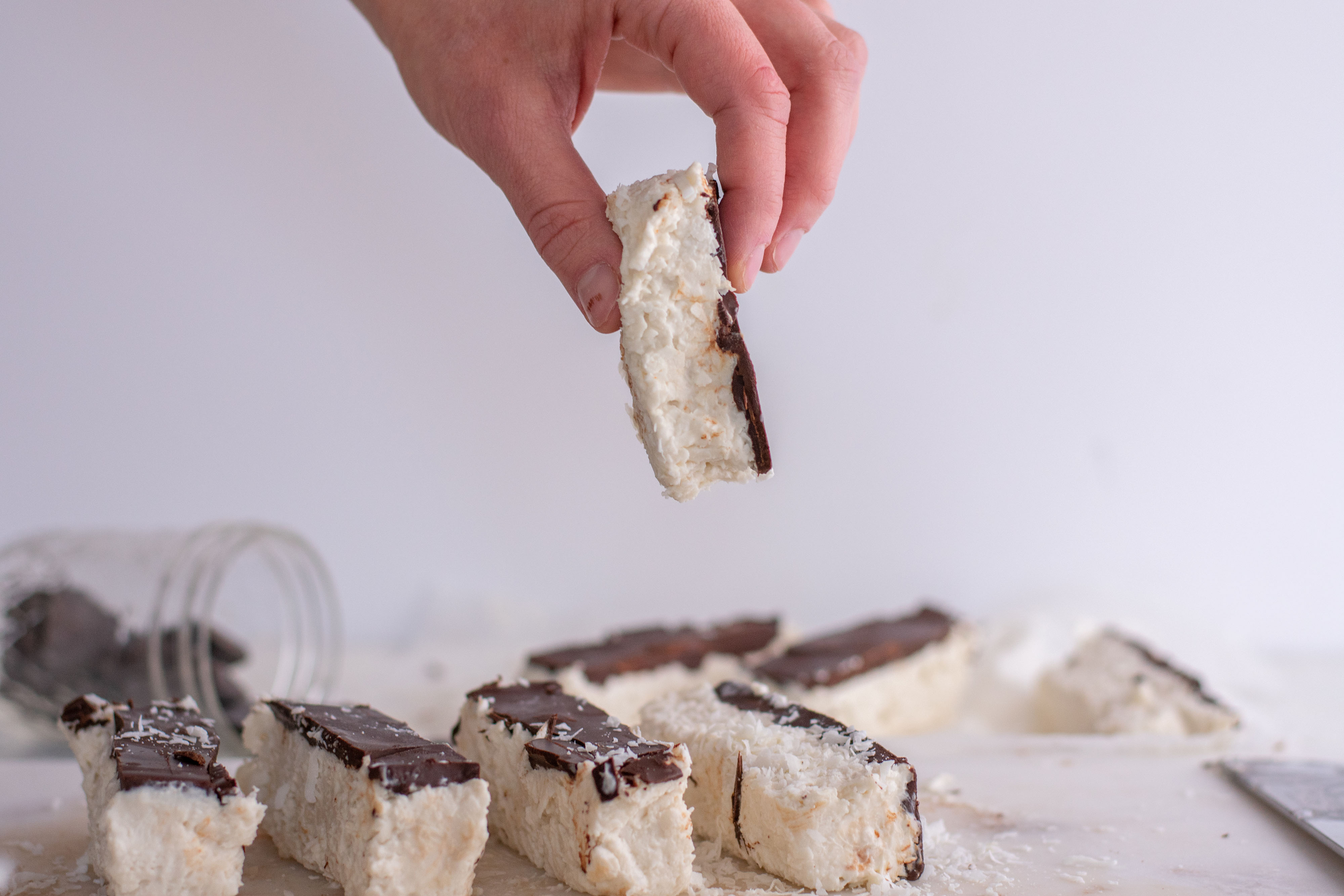 White coconut bars with a chocolate layer on top. laid out on a white surface with one picked up