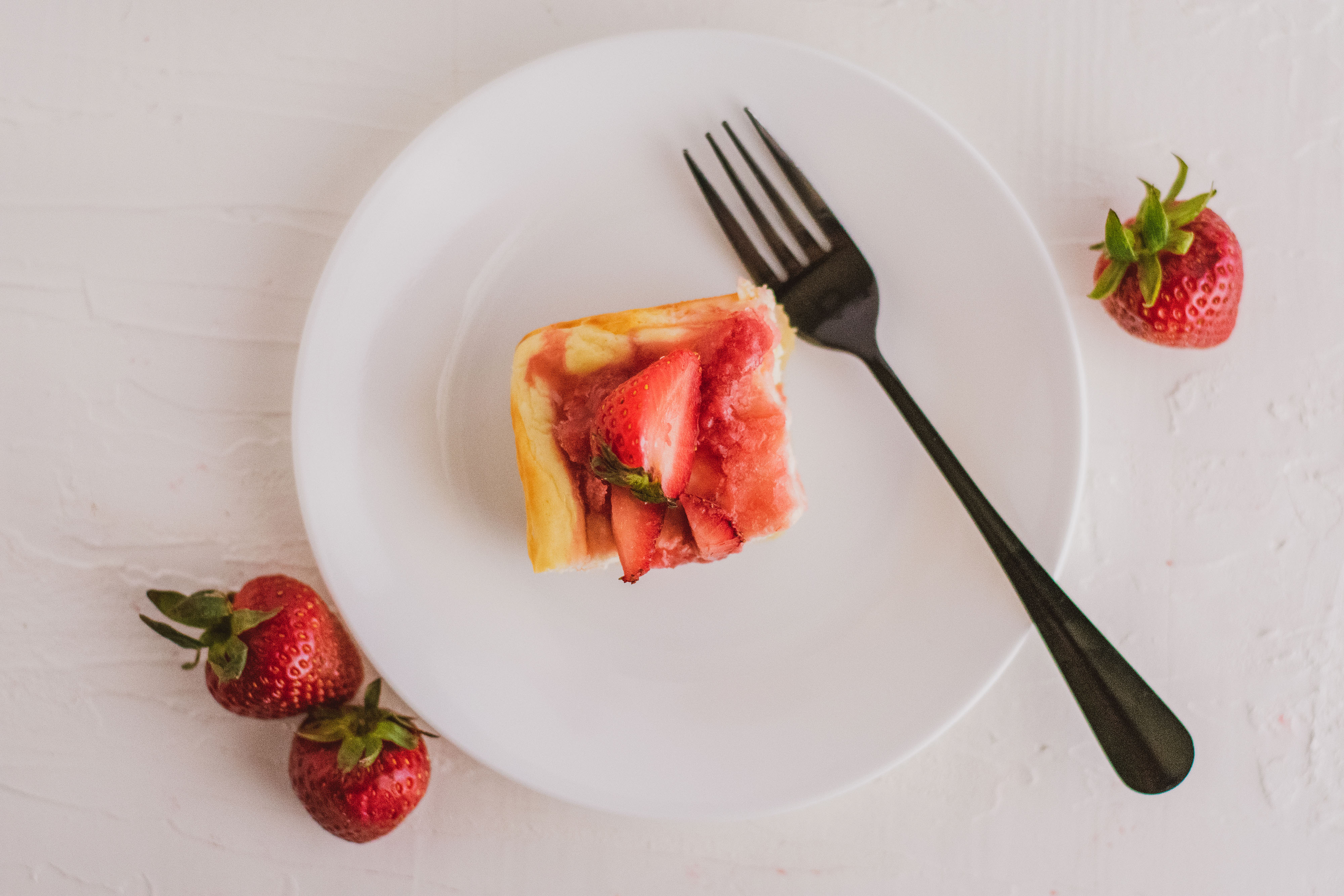 Low carb strawberry cheesecake with a strawberry jam on top on a white plate with a black fork on the side with strawberries on the sides. 