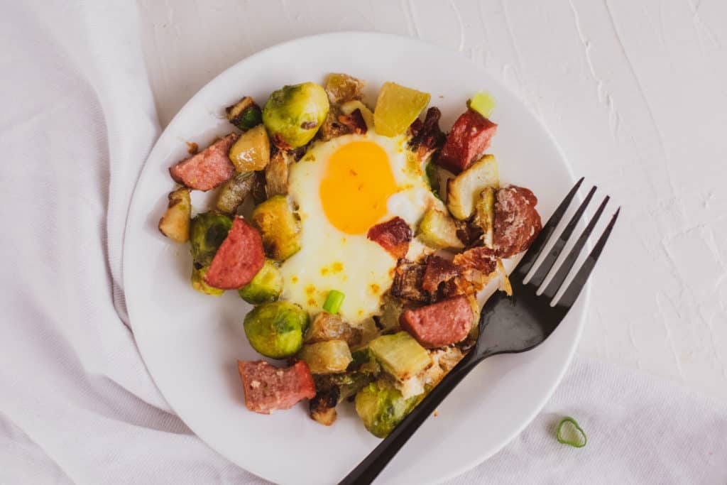 Keto Egg Bake on a white plate with a serving on a white surface. Loaded with sausage, Brussels sprouts and chayote.