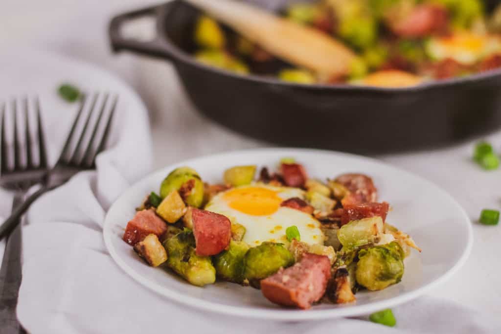 Keto Egg Bake in a cast iron skillet on a white surface. Loaded with sausage, Brussels sprouts and chayote and a white plate with a serving.