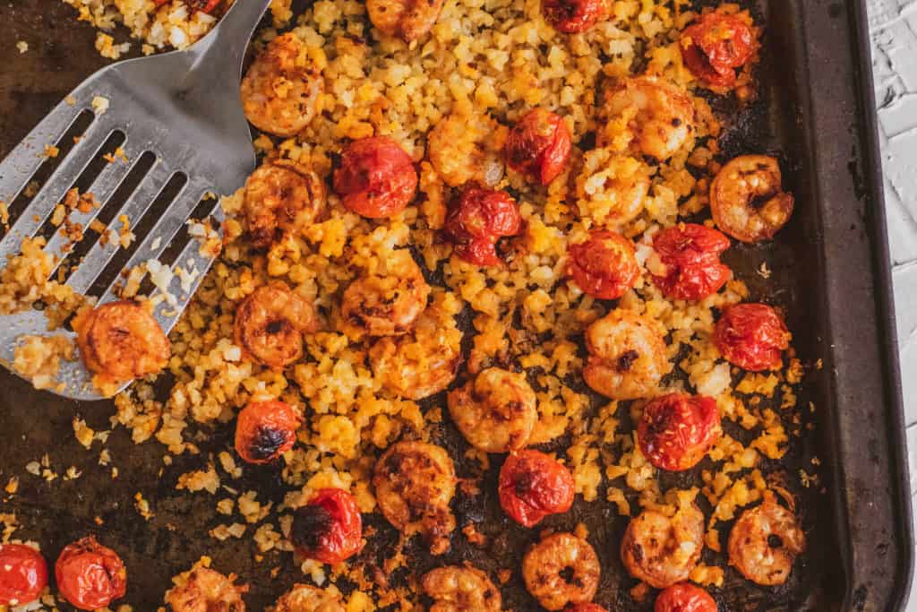 Keto Cajun Shrimp with roasted cherry tomatoes and cauliflower rice on a brownish black sheet pan and a metal spatula on the side..