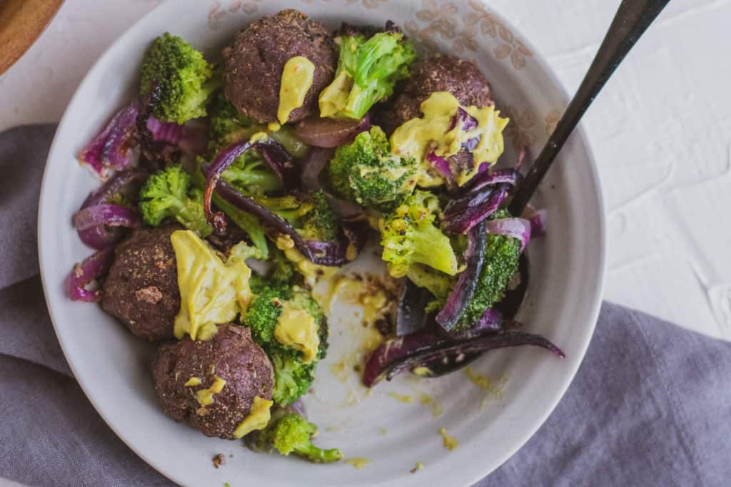 Low carb keto meatballs with a creamy avocado sauce in a bowl on a white surface with a black spoon and roasted broccoli and red onions