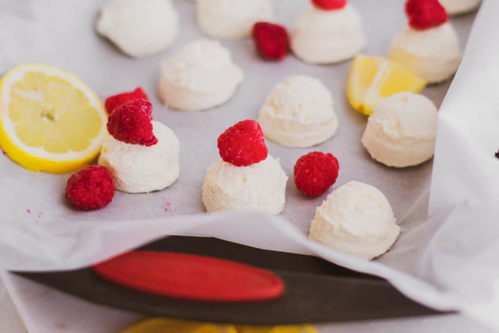 Keto creamy lemon fat bombs with raspberries and lemon slices on a sheet pan with parchment paper