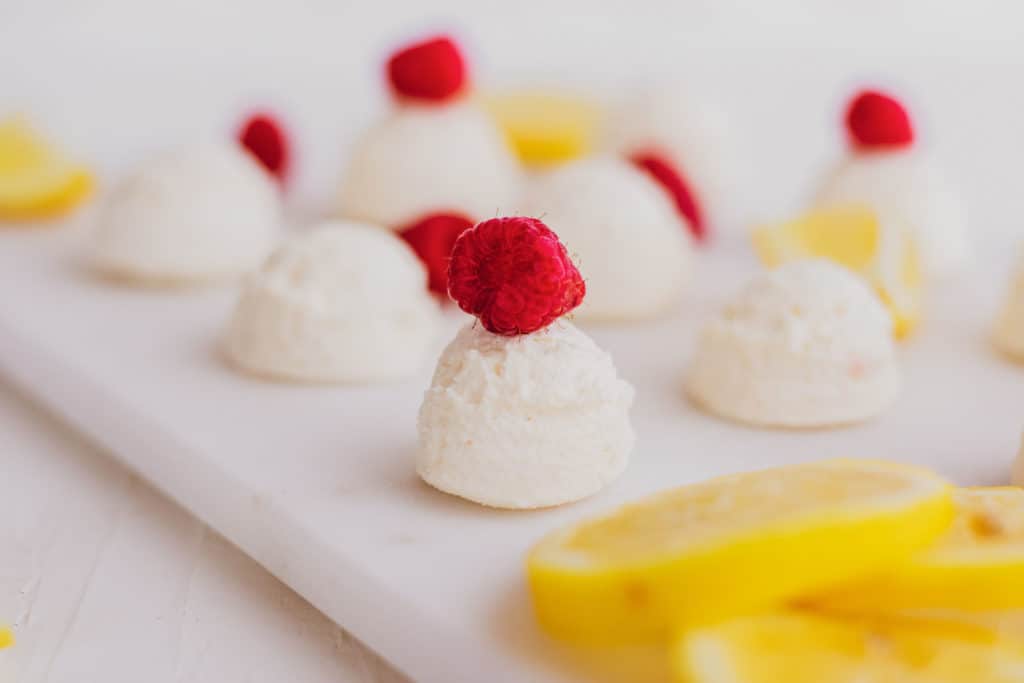 Keto creamy lemon fat bombs with raspberries and lemon slices on a white surface