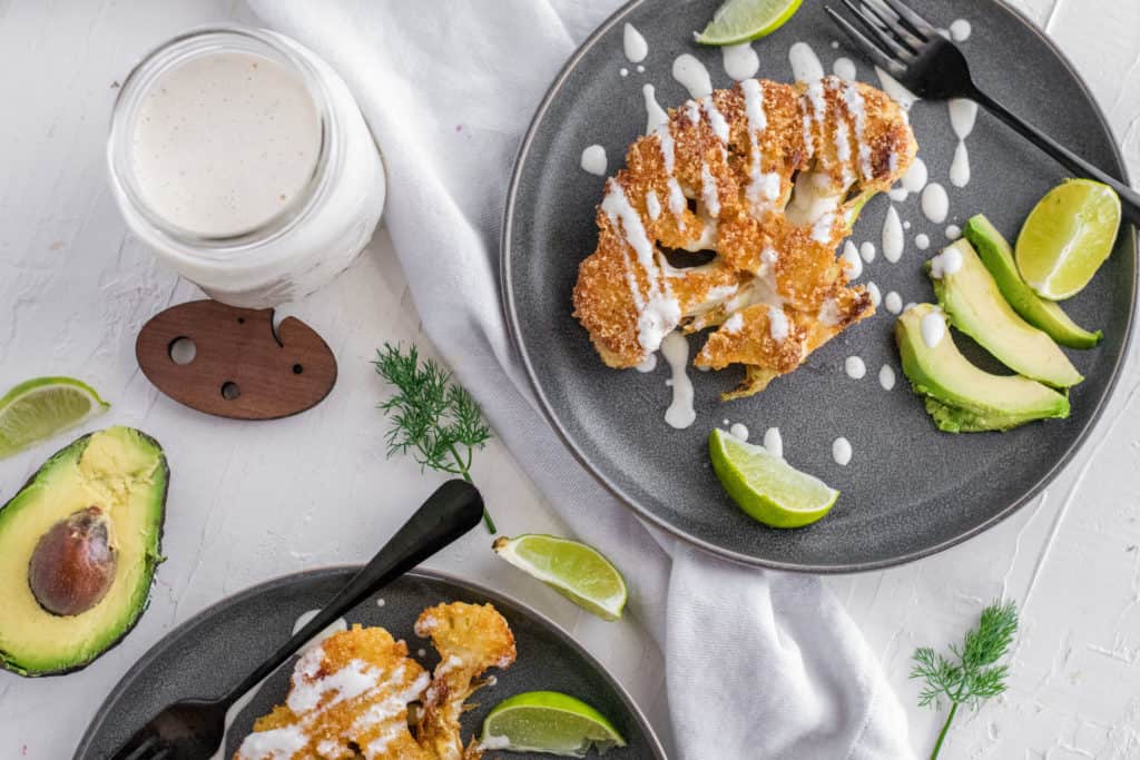 Fried pork rind covered cauliflower steaks on a grey plate with ranch on top and avocado and limes on the side.