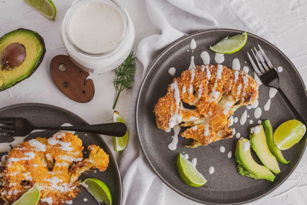 Fried pork rind covered cauliflower steaks on a grey plate with ranch on top and avocado and limes on the side.