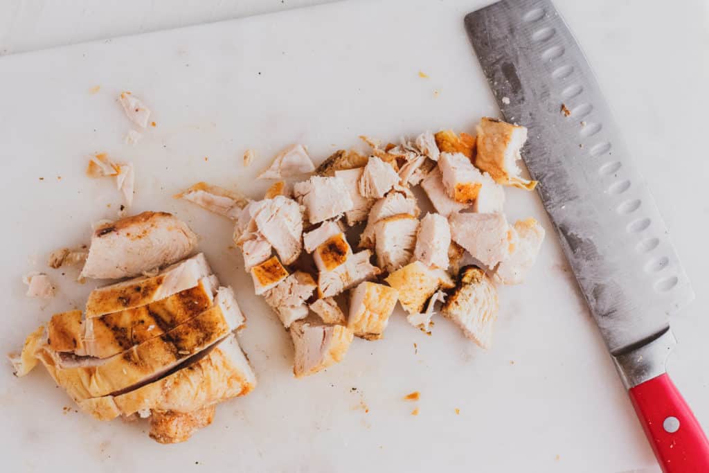 A grilled chicken breast chopped with a butcher knife on the side on a white surface.