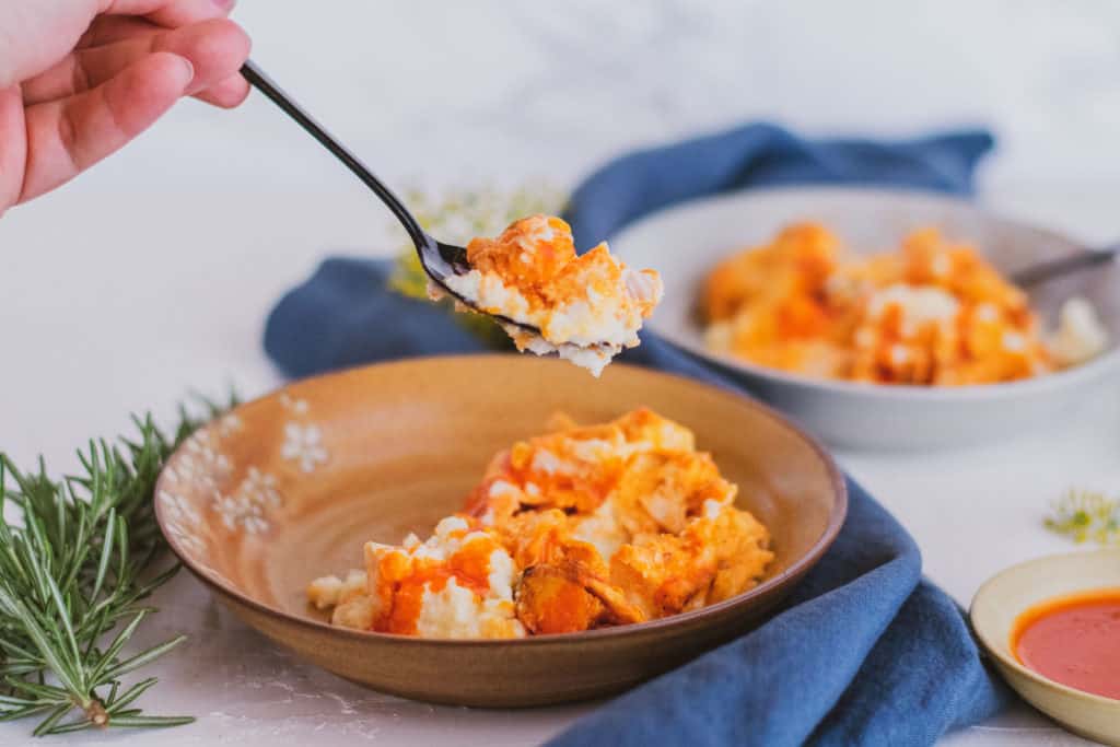 Low carb mashed cauliflower buffalo chicken in a bowl on a white surface with a blue napkin on the side with a black spoon taking a bite.