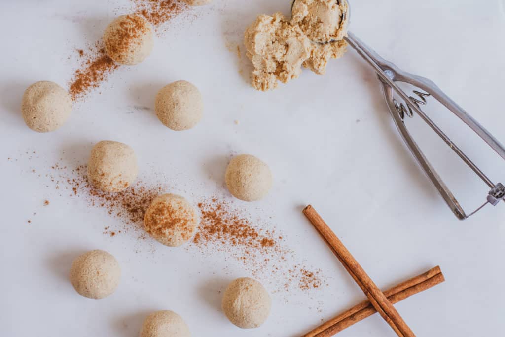 Keto Pumpkin Spice Fat Bombs rolled into balls with cinnamon on top on a white surface with cinnamon sticks and a cookie scoop on the side