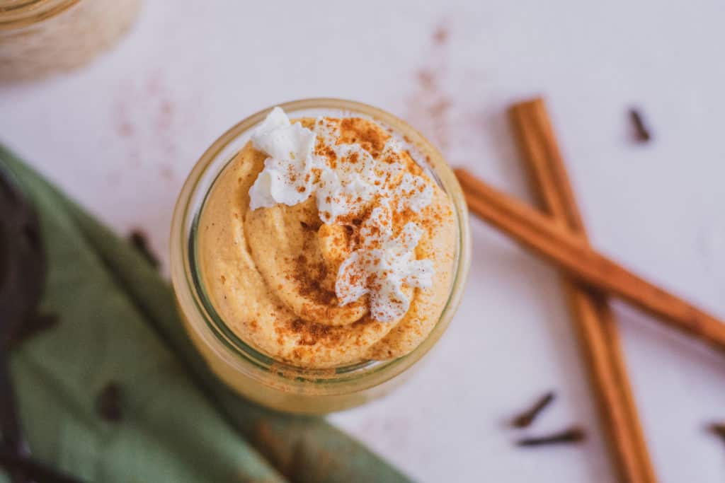 Low carb pumpkin cheesecake mousse in a clear jar with cinnamon sticks, black spoons, and a green linen on the side. Keto dessert.