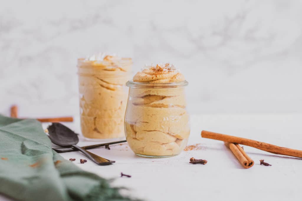 Low carb pumpkin cheesecake mousse in a clear jar with cinnamon sticks, black spoons, and a green linen on the side. Keto dessert.