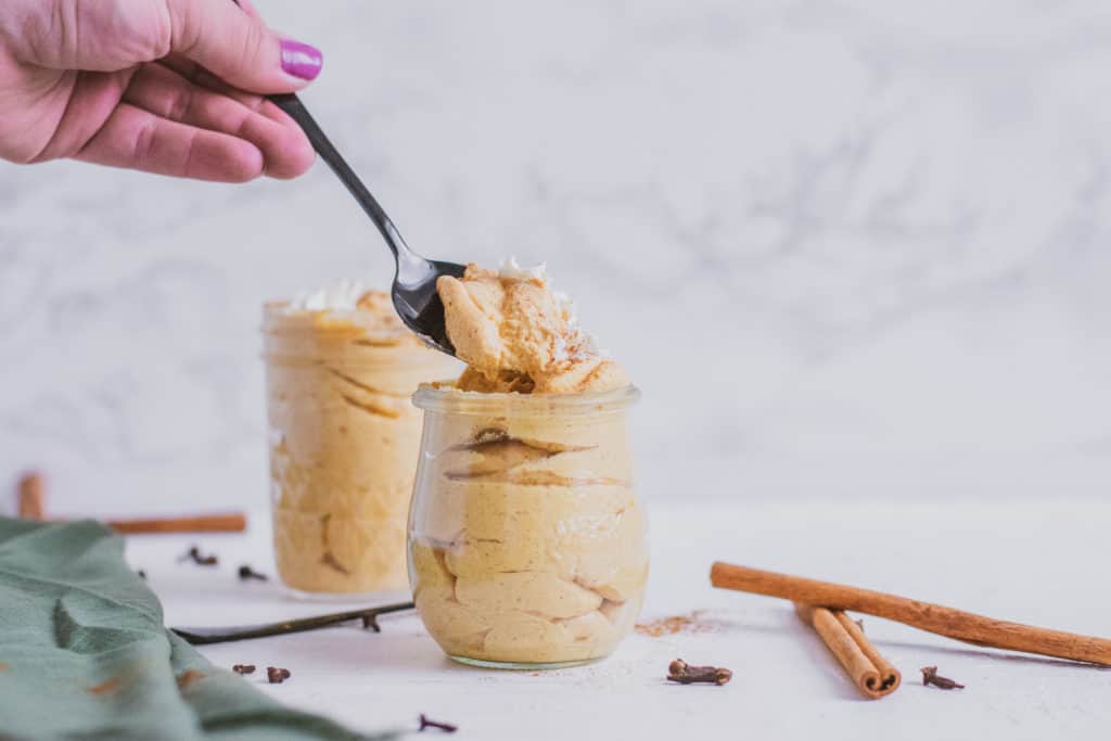 Low carb pumpkin cheesecake mousse in a clear jar with cinnamon sticks, black spoons, and a green linen on the side with a spoon fell being taken out. Keto dessert.