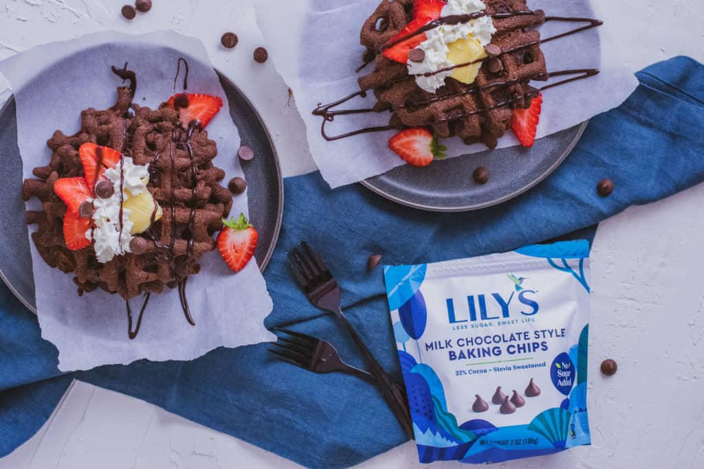 Delicious low carb Keto Chocolate Waffles on a plate topped with butter, strawberries, cream and chocolate syrup with a blue napkin on the side and a bag of lilys chocolate chips.