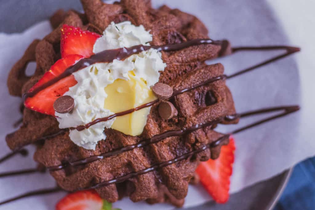 Delicious low carb Keto Chocolate Waffles on a plate topped with butter, strawberries, cream and chocolate syrup with a blue napkin on the side.