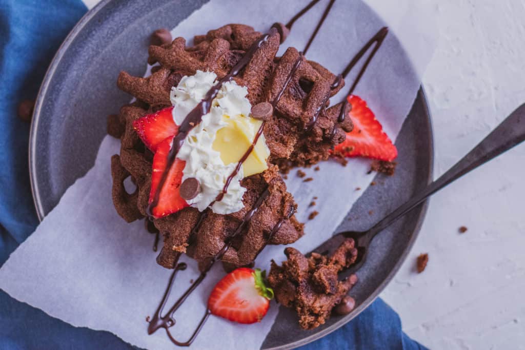 Delicious low carb Keto Chocolate Waffles on a plate topped with butter, strawberries, cream and chocolate syrup with a blue napkin on the side.