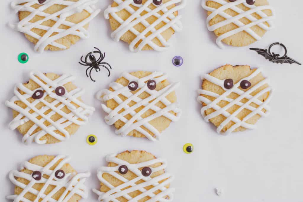 Keto Mummy sugar cookies with cream cheese frosting lines on the cookies and chocolate chip eyes on a white surface.
