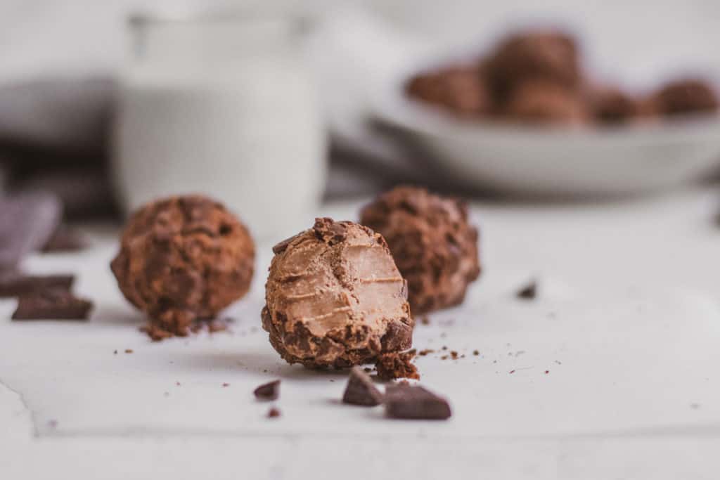 Keto peanut butter brownie fat bombs with a bite taken out on a white surface with a glass of almond milk in the background and a grey napkin and a bowl of the recipe.