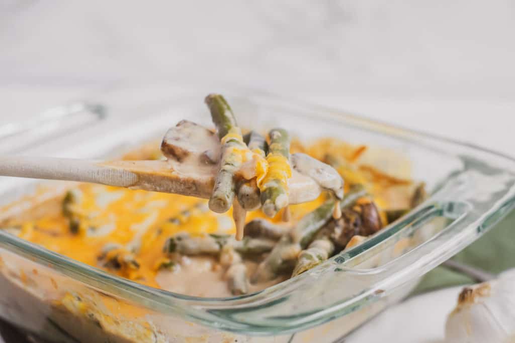 Low carb green bean casserole with melted cheese on top in a casserole dish with a wooden spoon picking up a serving.