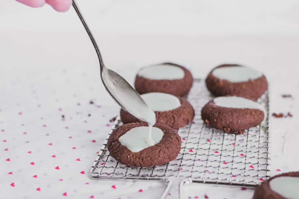 Keto Chocolate Thumbprint Cookies with white chocolate ganache in the middle on a wire wrack and heart paper on the bottom on a white surface.