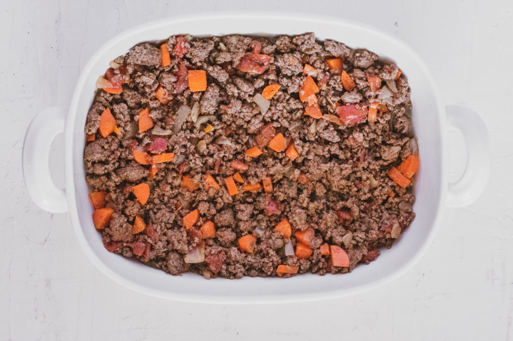 Ground beef layer in a white casserole dish on a white surface.