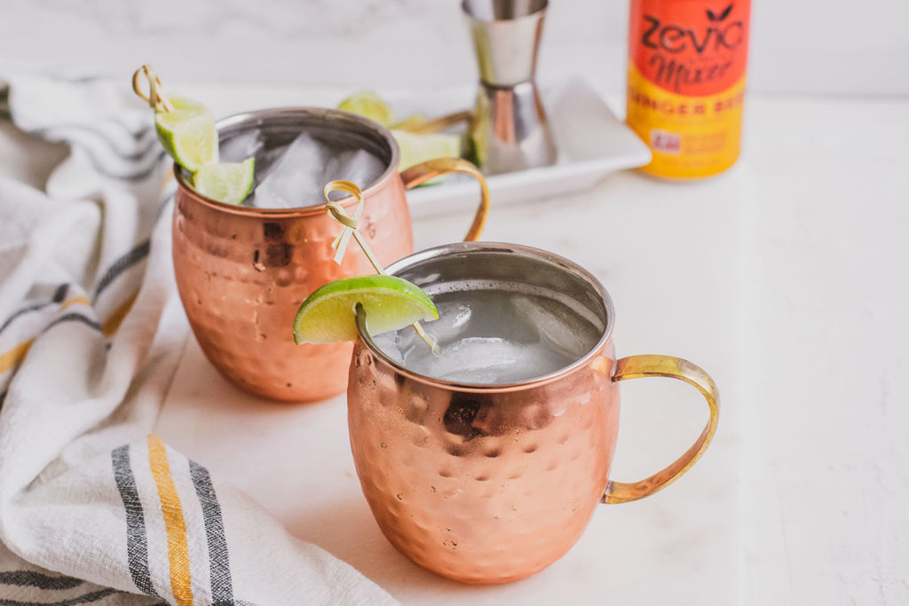 Keto Moscow Mule in brass mugs with a lime slice on the rim on a white surface and a Zevia ginger beer mixer on the side.
