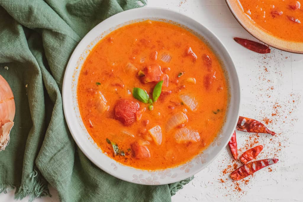 Creamy Moroccan Tomato Soup in a white bowl on a white surface with red chiles on the side and a green napkin.