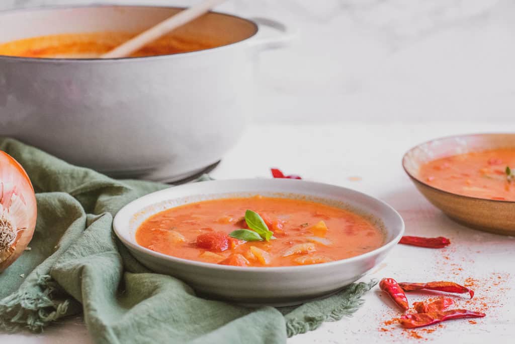 Creamy Moroccan Tomato Soup in a white bowl on a white surface with red chiles on the side and a green napkin.