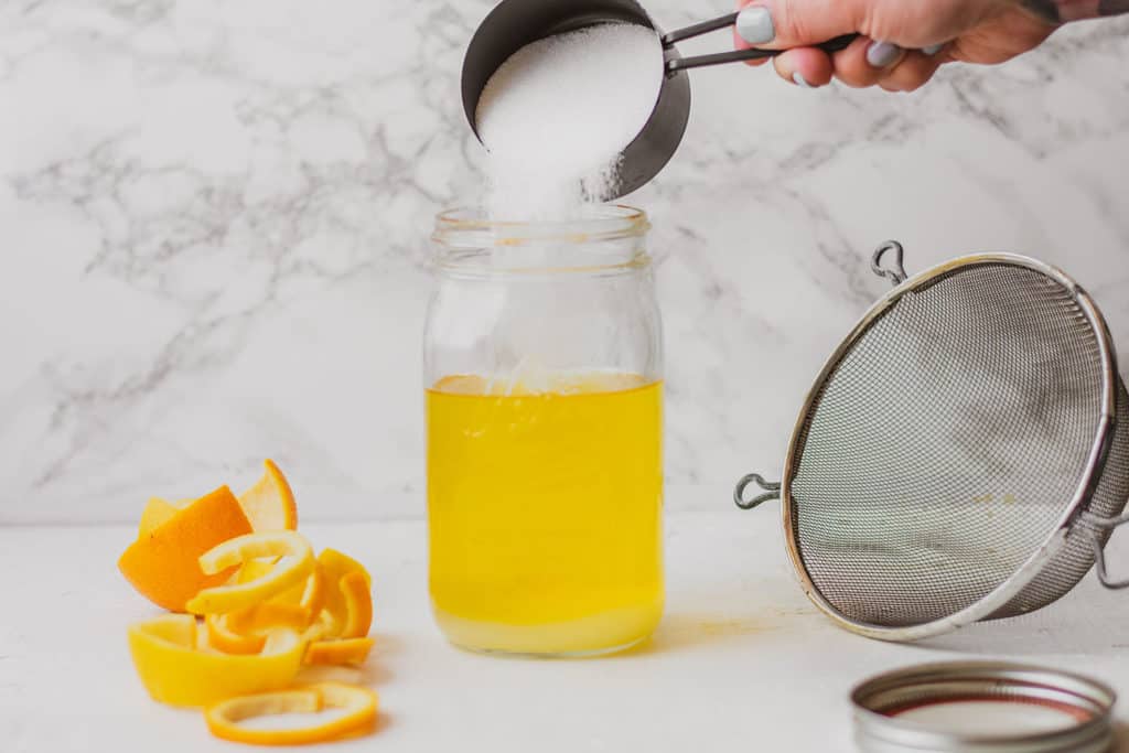Keto Triple Sec in a mason jar with erythritol pouring in the jar on a white surface with oranges and lemons on the side.