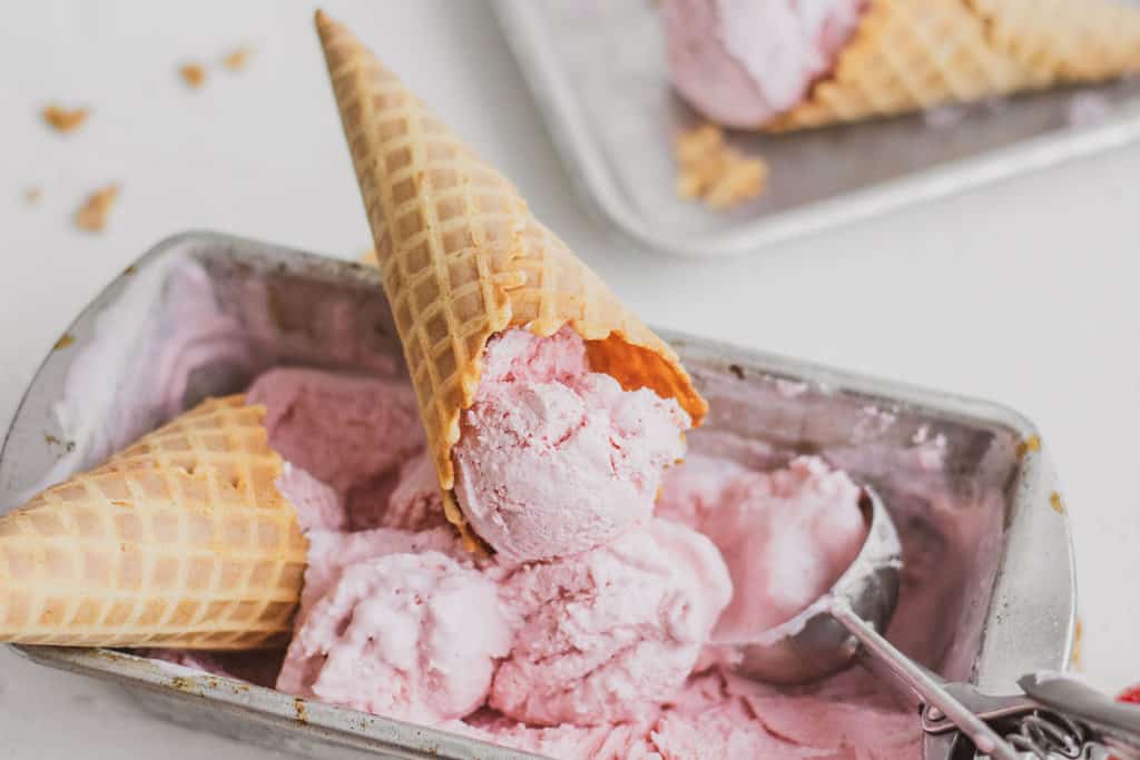 Keto strawberry ice cream in a tin bread pan in a waffle cone on a white surface.