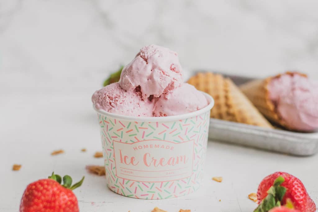 Keto strawberry ice cream in a ice cream bowl whit ice cream in a waffle cone in the background on a white surface.