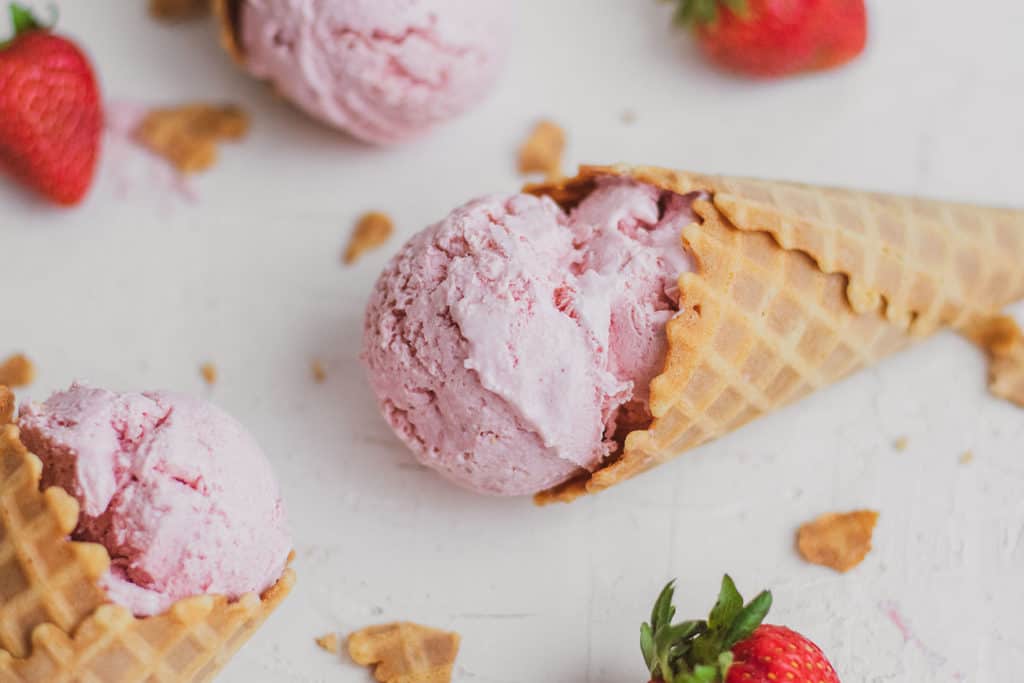Keto strawberry ice cream in a waffle cone on a white surface.