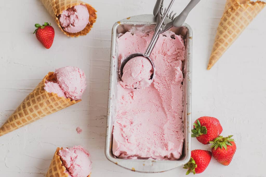 Keto strawberry ice cream in a tin bread pan in a waffle cone on a white surface.