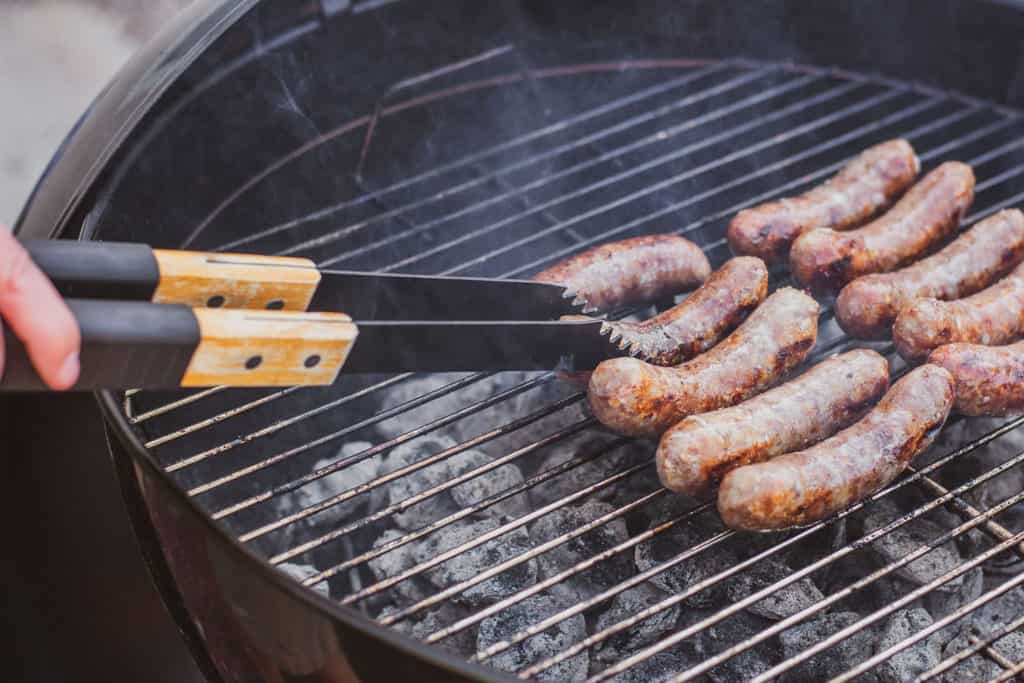 Low carb Wisconsin Beer Brats on a Weber grill with a woman flipping the brats. 