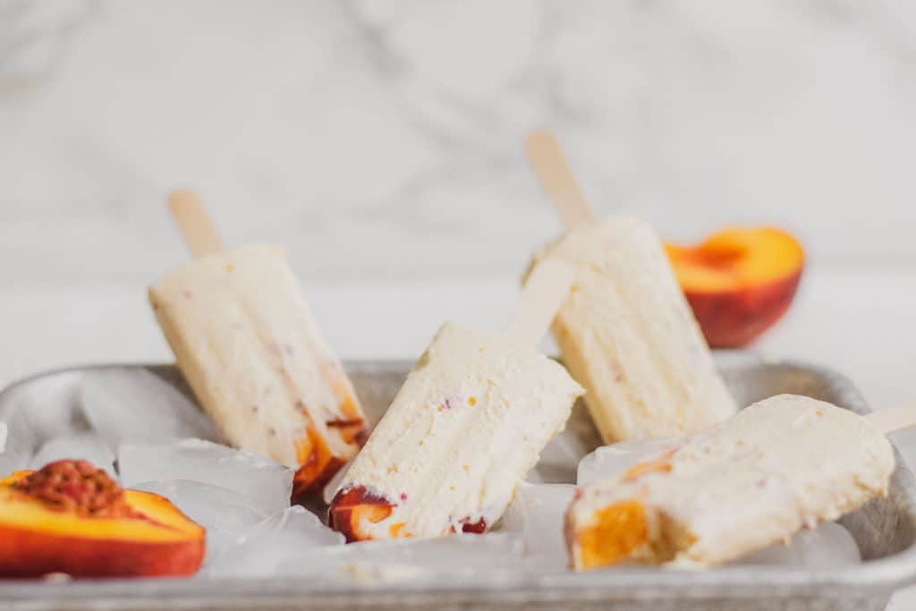 Keto peach ice cream bars on a metal tray with ice and fresh peaches.