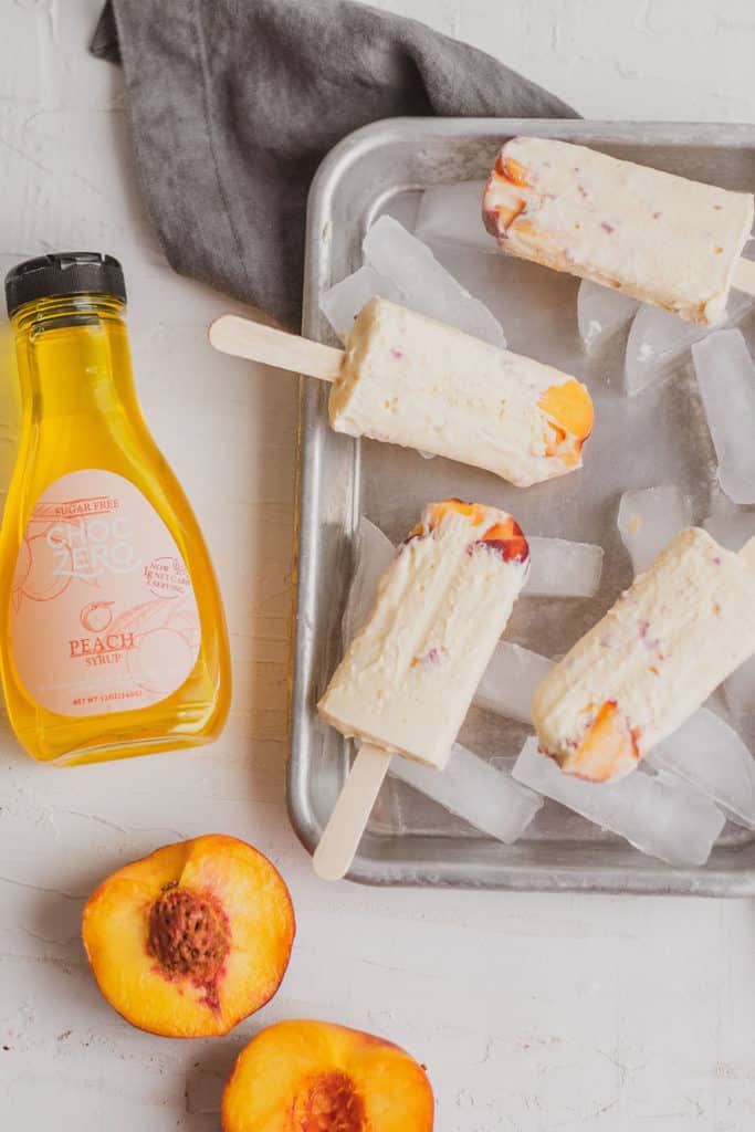 Keto peach ice cream bars on a metal tray with ice and fresh peaches and a bottle of ChocZero Peach Syrup.