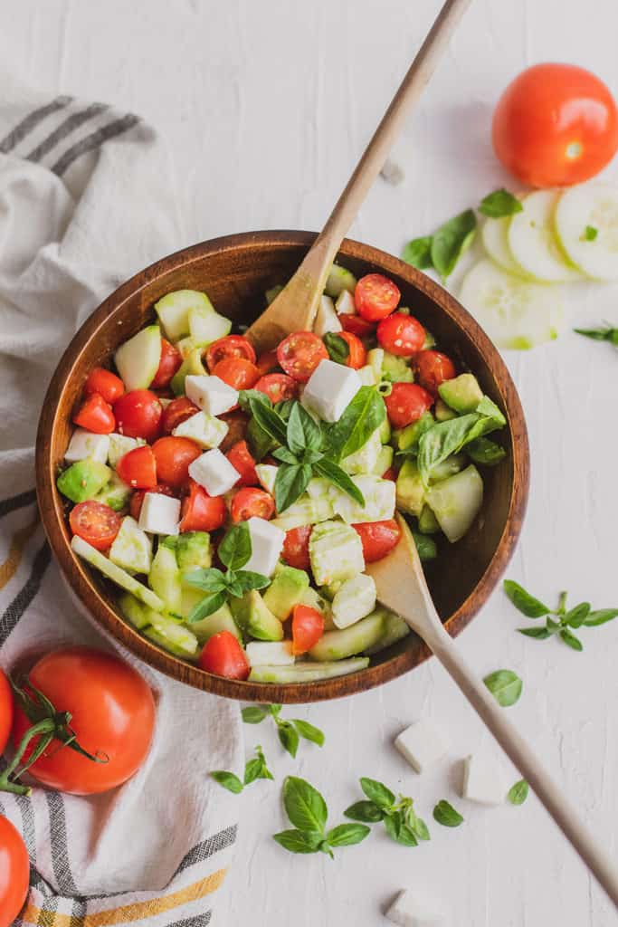 Fresh keto Tomato Cucumber Salad in a wood bowl on a white surface with wood spoons.
