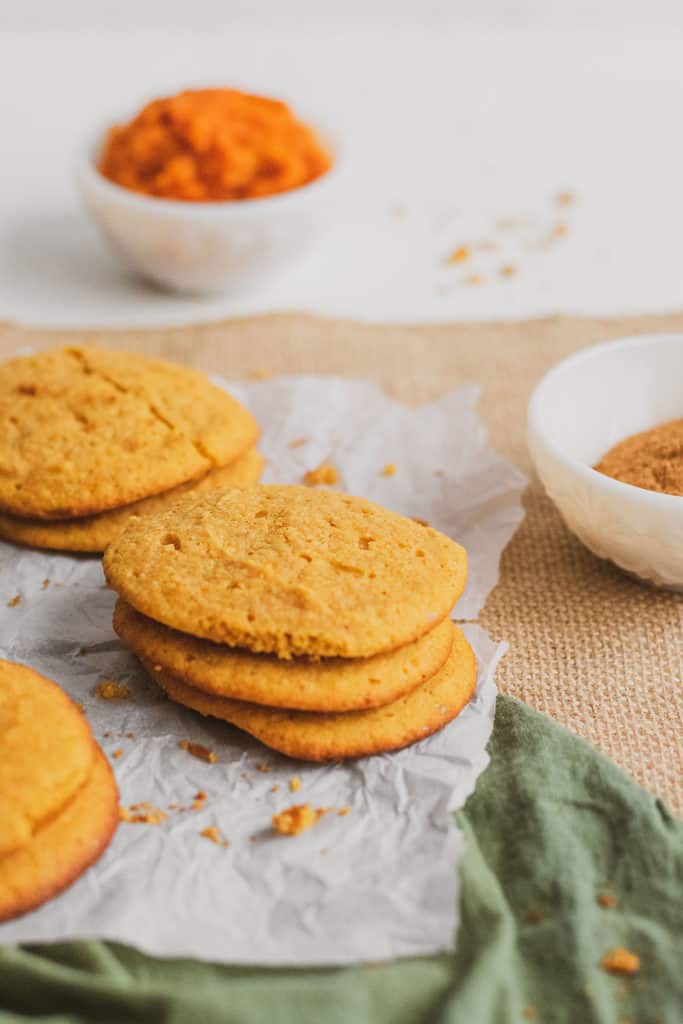 Keto pumpkin cookies stacked on parchment paper with ingredients on the side